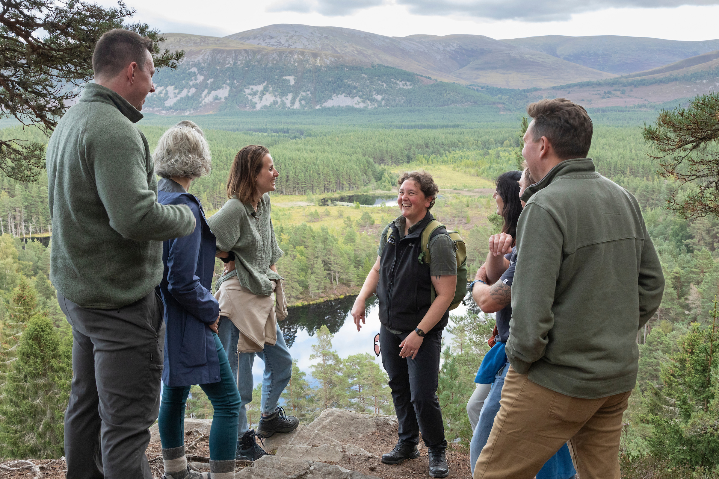 A group of six people stand on a rocky overlook, engaged in conversation and smiling. They are surrounded by a forest with a small lochan below, and d