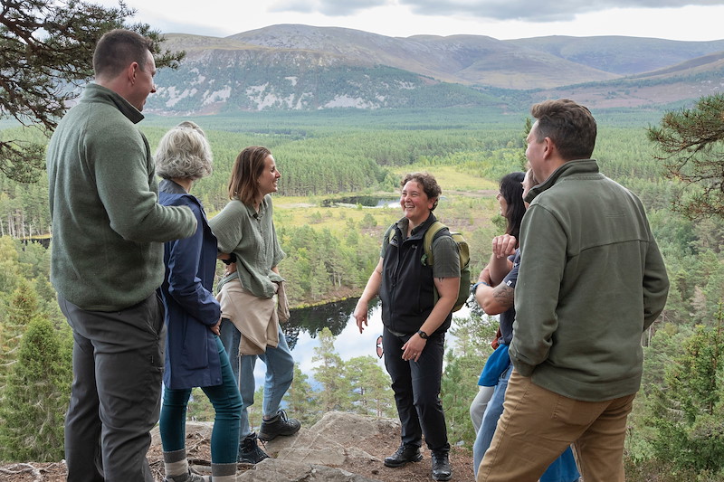 Rewilding Journeys guide, Stef Lauer, talking to guests at a viewpoint in the forest overlooking Uath Lochans in the Cairngorms National Park A group of six people stand on a rocky overlook, engaged in conversation and smiling. They are surrounded by a forest with a small lochan below, and d